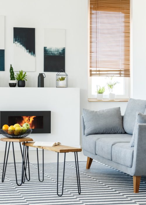 Real photo of a grey couch standing in front of a small wooden table in living room interior with a fireplace and posters on walls